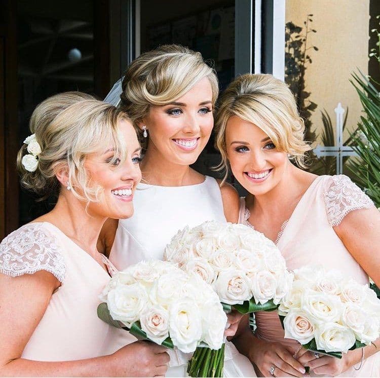 Bride and two bridesmaids with their hair pinned up, holding bouquets of white roses