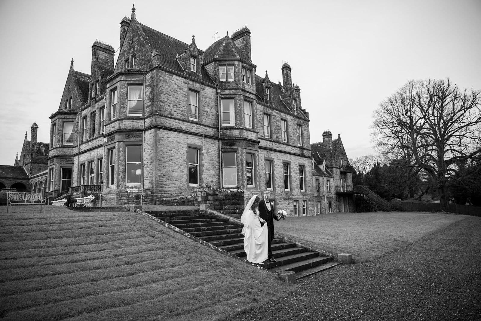 Bride and groom on the steps of a large stone house in winter, in black and white