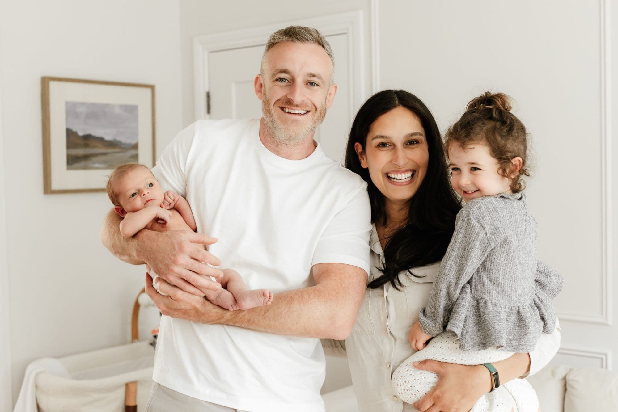 Smiling parents in a white panelled room, he holding a newborn and she a toddler