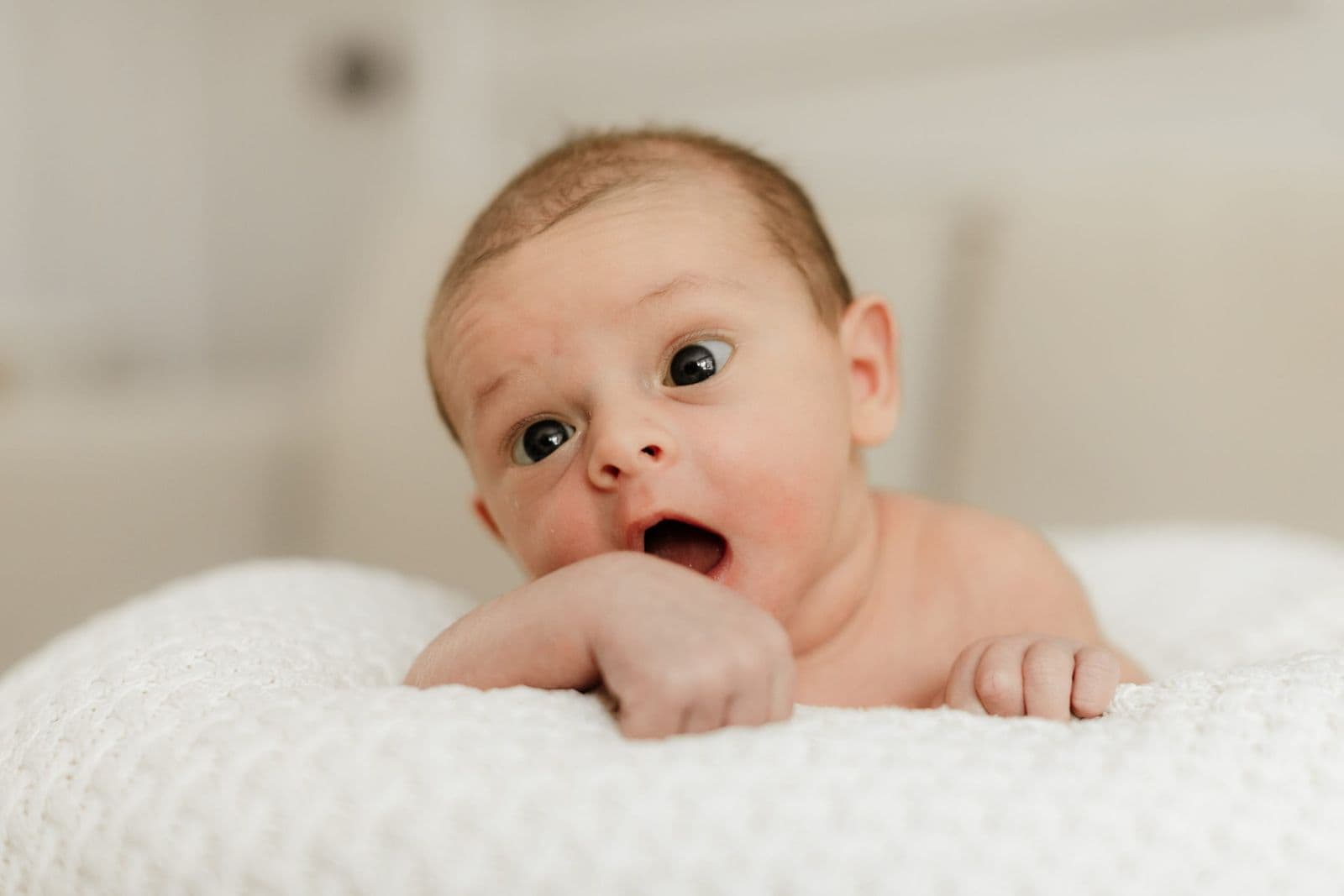 Wide eyed newborn on its front on a white knitted blanket, mouth open