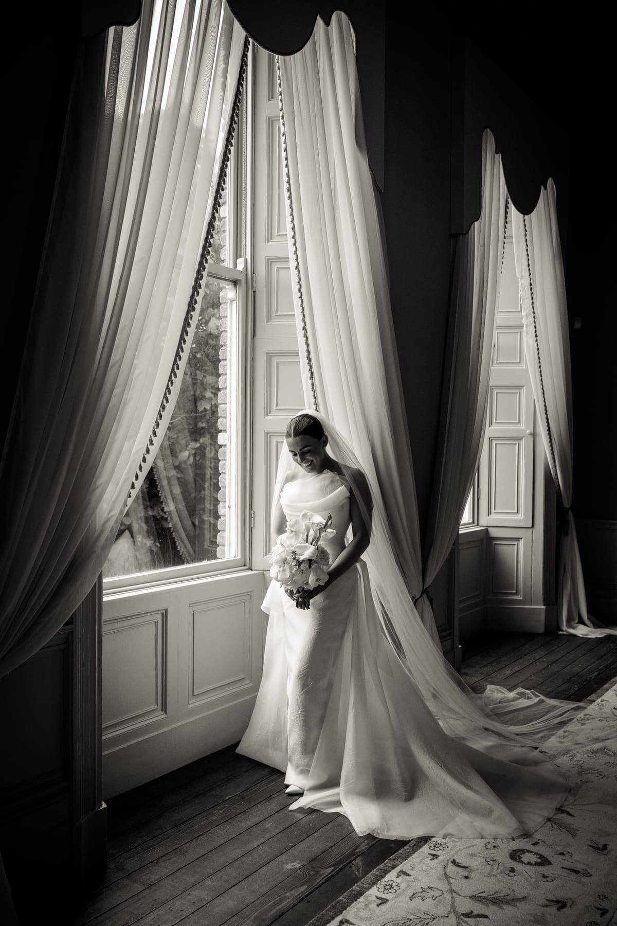 Bride at a window between long curtains, in black and white