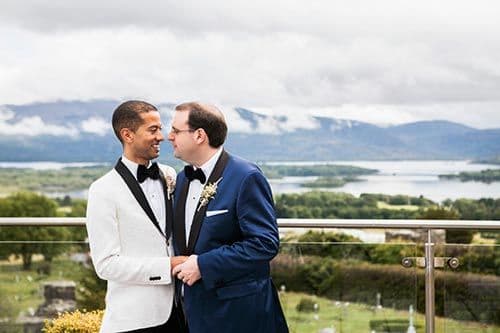 Two grooms laughing together on a balcony above a lake