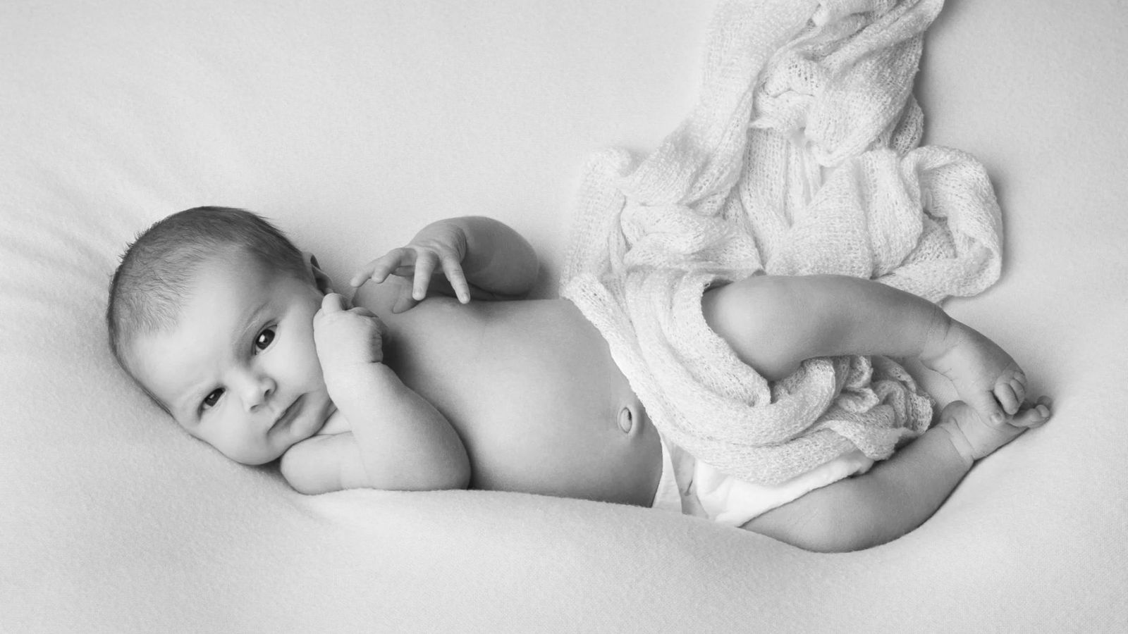 Baby lying on its side on a white blanket, looking at the camera, in black and white