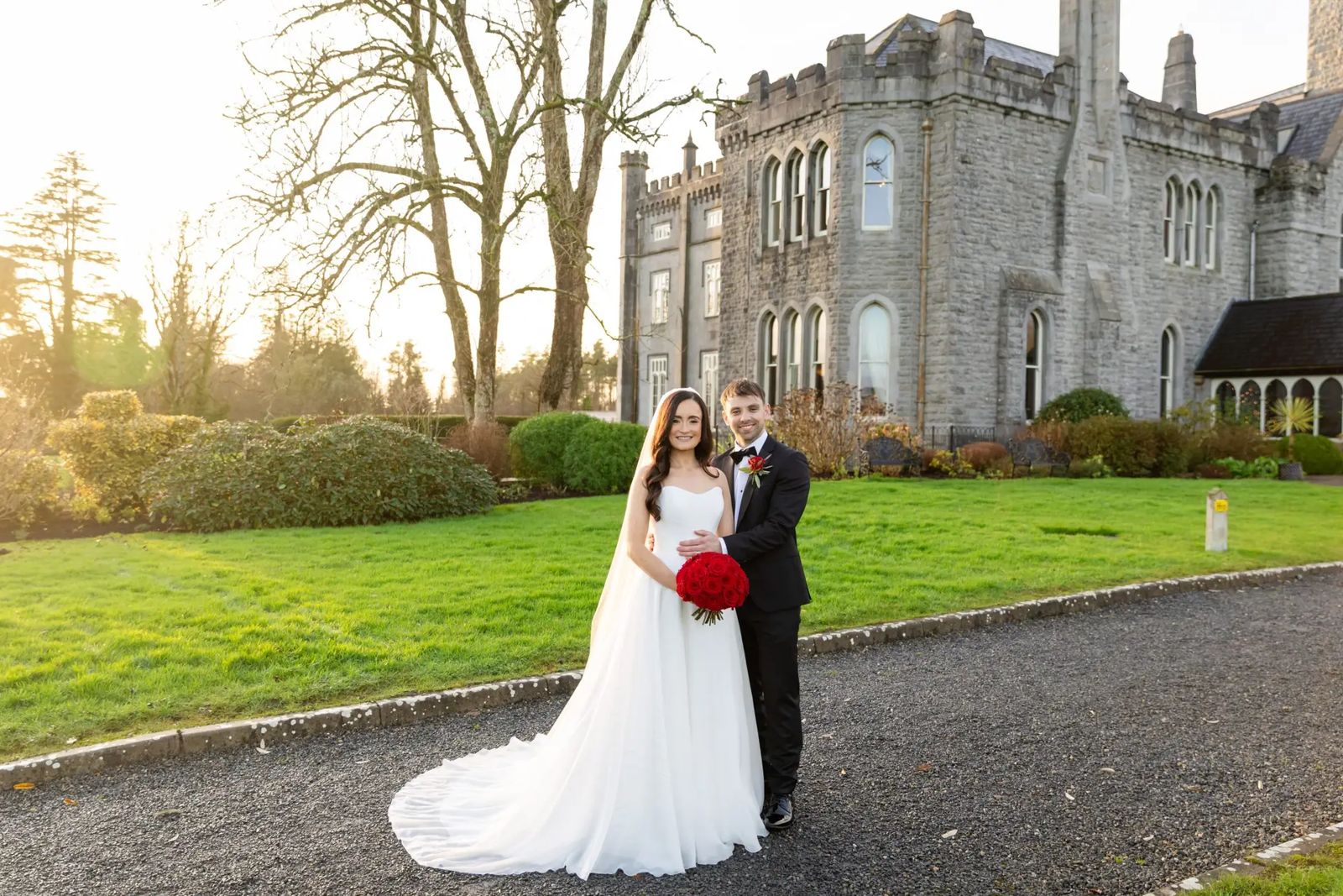 Bride with a bouquet of red roses and her groom on the drive of a grey stone castle, in low sun