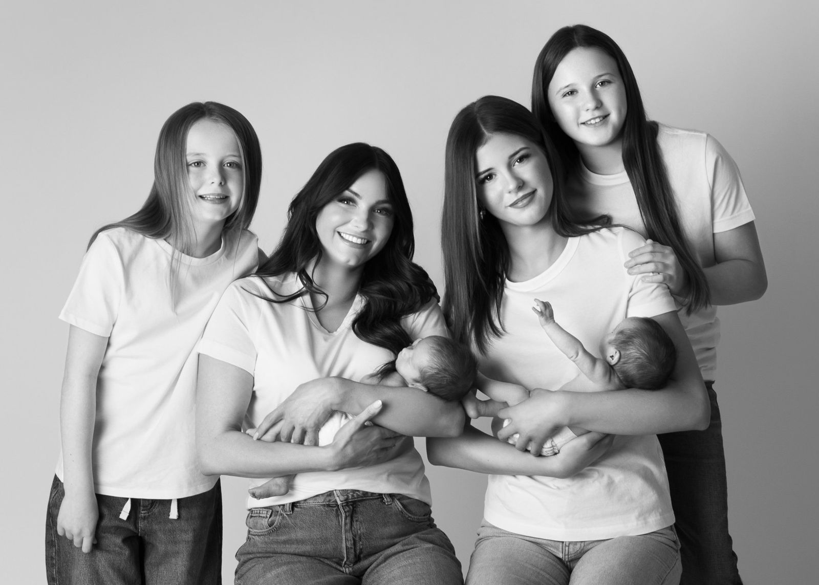 Two young women in white T-shirts each holding a newborn baby, with two girls leaning in beside them, in black and white