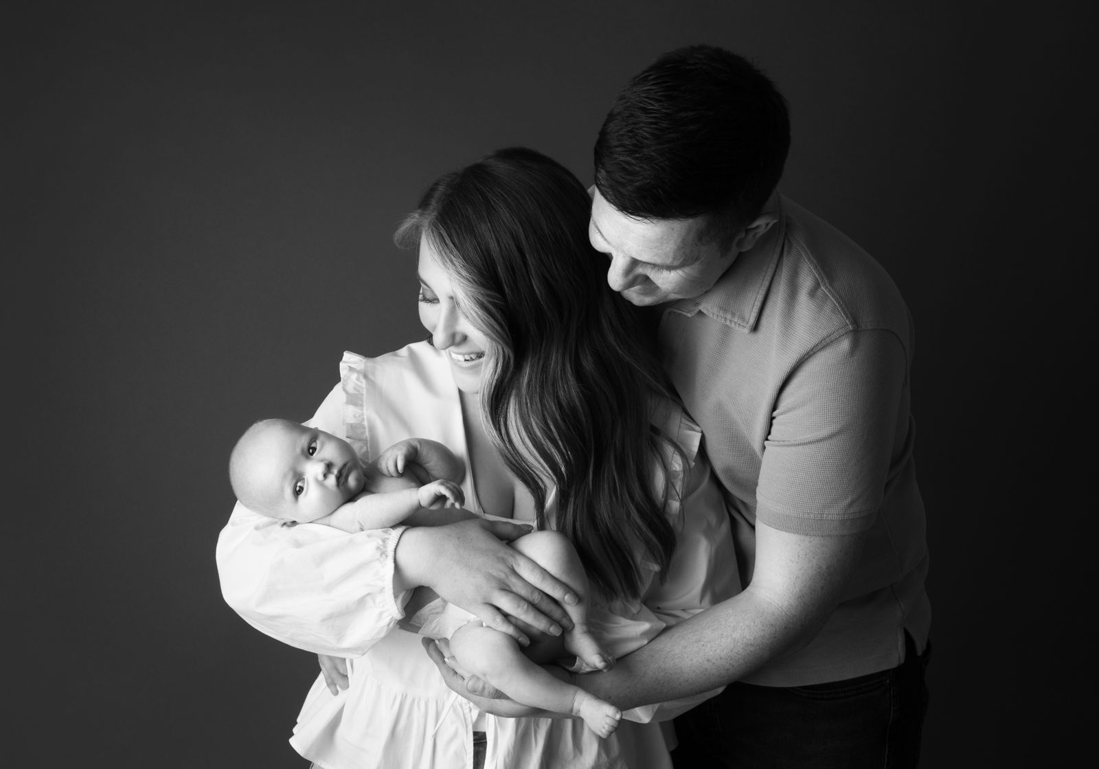 Mother holding her baby, who looks at the camera, with the father's arms around them both, in black and white